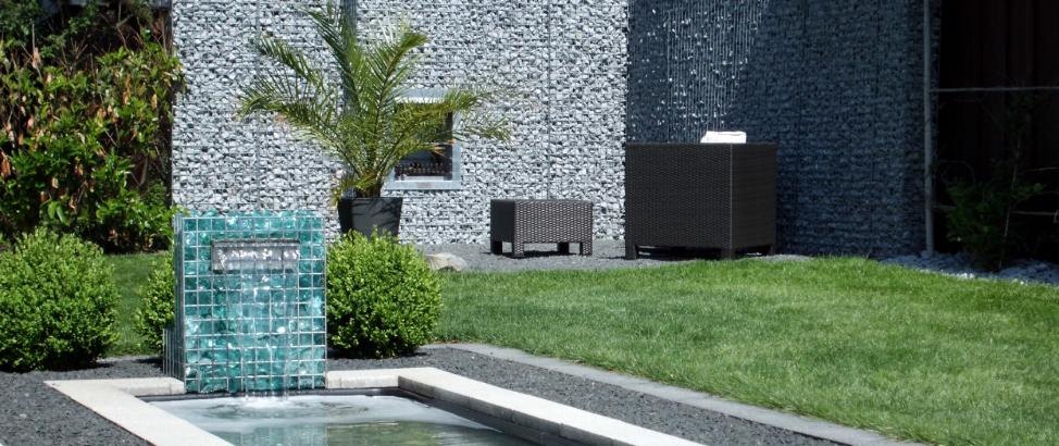 Ein moderner Garten mit Gabionen-Gartenkamin und Wasserfall, kombiniert mit einem elegant gestalteten Teich und Gartenmöbeln